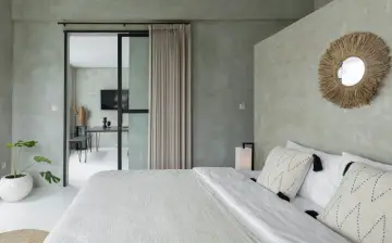 Ett modernt grått sovrum med en stor säng, en dörröppning till ett annat rum med ett skrivbord och en TV, samt en dekorativ spegel på väggen.
