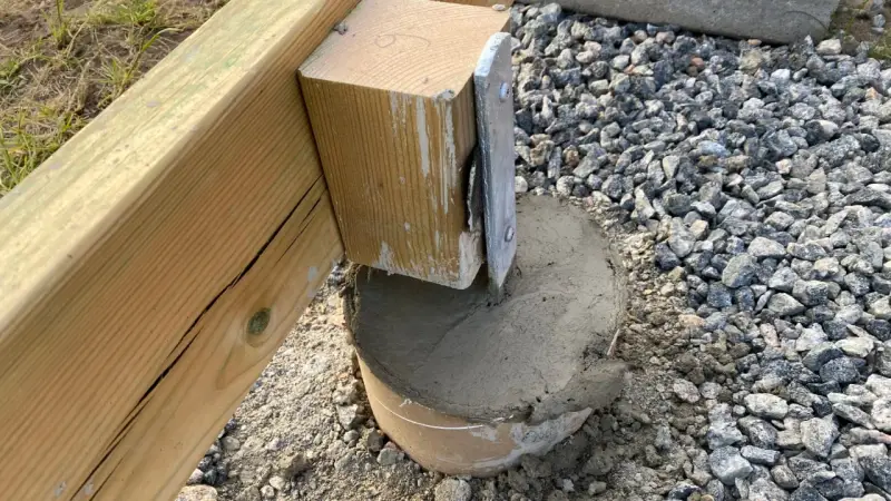 En trästolpe fästs i en betongplint, en del av arbetet med att bygga altan på plintar. Grus och jord syns runt plinten.